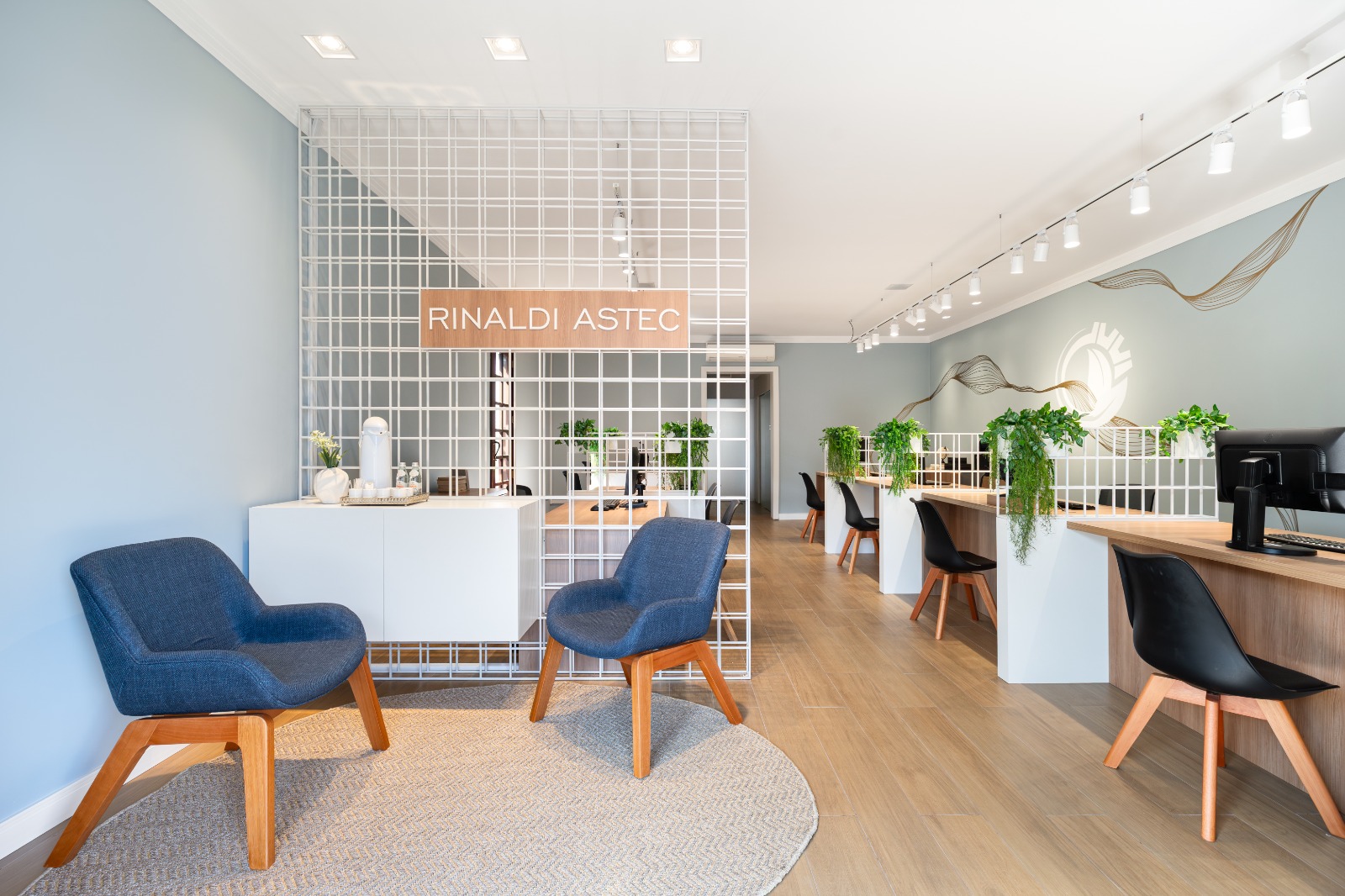 Recepção da Rinaldi Astec, com poltronas, a grade branca com a placa e as mesas da equipe